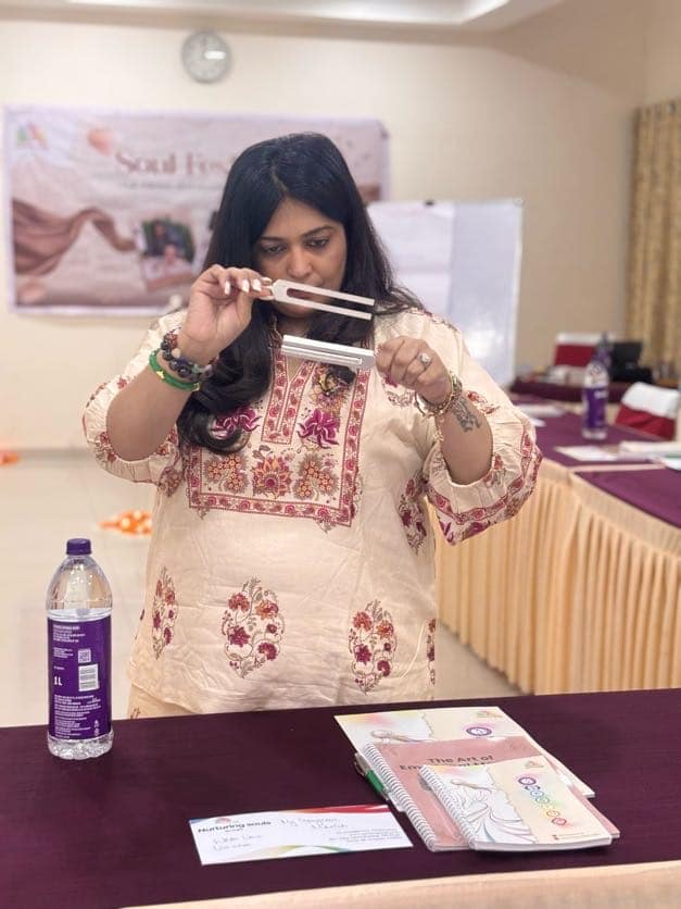 Megha holding a struck tuning fork in front of a Soul Festival banner, a workbook open on the table beside her
