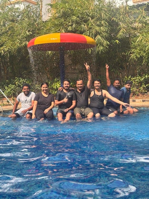 Participants sitting together on the edge of a swimming pool under a bright parasol fountain, feet in the water