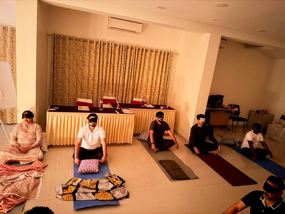 A group lying and sitting on mats across a hall floor during a guided meditation, the facilitator seated at the front