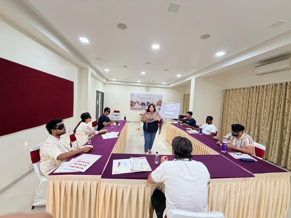 Participants seated along long tables writing in workbooks while a facilitator leads from the front of the hall